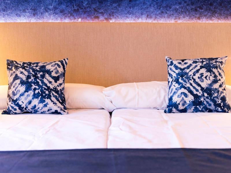 Zwei weiße Betten mit beigen Kopfteil und zwei blauen Kissen mit Muster darauf.
