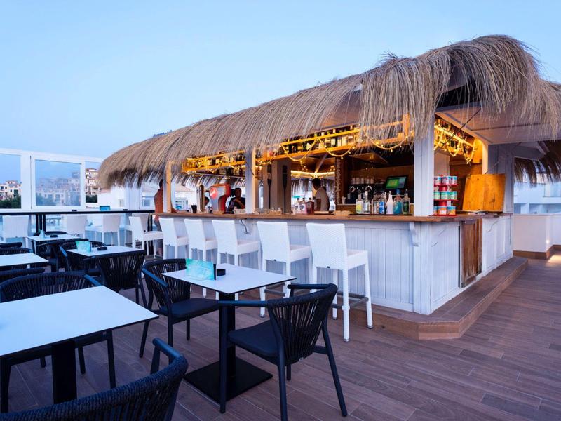 Offene Strandbar mit Strohdach, weißen Tischen und schwarzen Stühlen auf Holzterrasse bei Abendlicht.