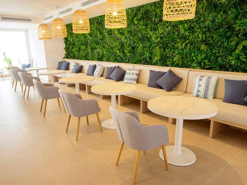 Moderner Gastraum mit hellen Tischen, grauen Stühlen und einer Wand mit grünem Pflanzenrelief.