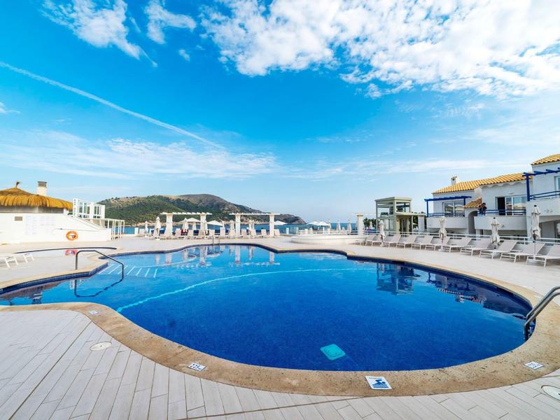Schwimmbad mit blauem Wasser, umgeben von weißem Boden und Liegestühlen unter blauem Himmel.
