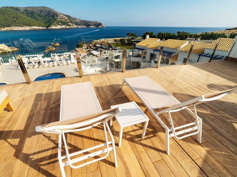 Drei weiße Liegestühle und ein Tisch auf hellbraunem Holzdeck mit Blick auf das blaue Meer und Hügel.