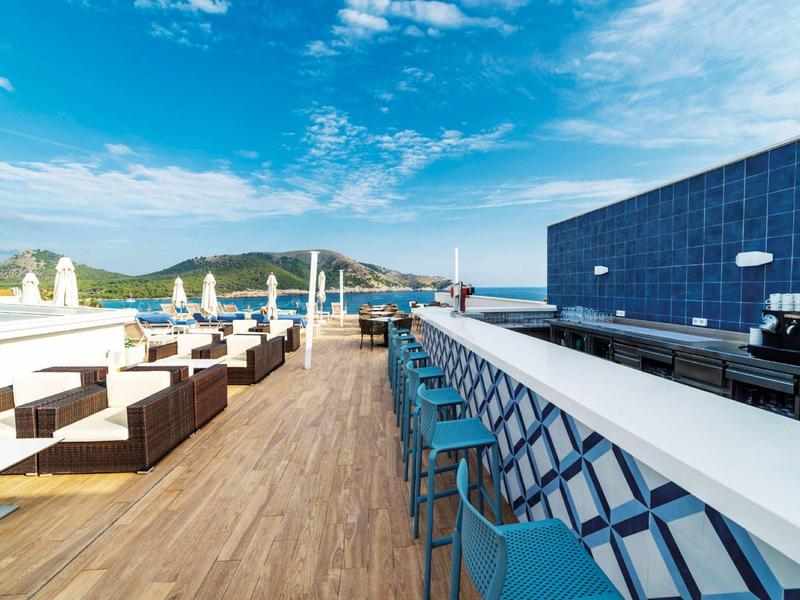 Moderne Dachterrasse mit blauen Stühlen, weißen Sonnenliegen und weiter Aussicht auf Himmel und Hügel.