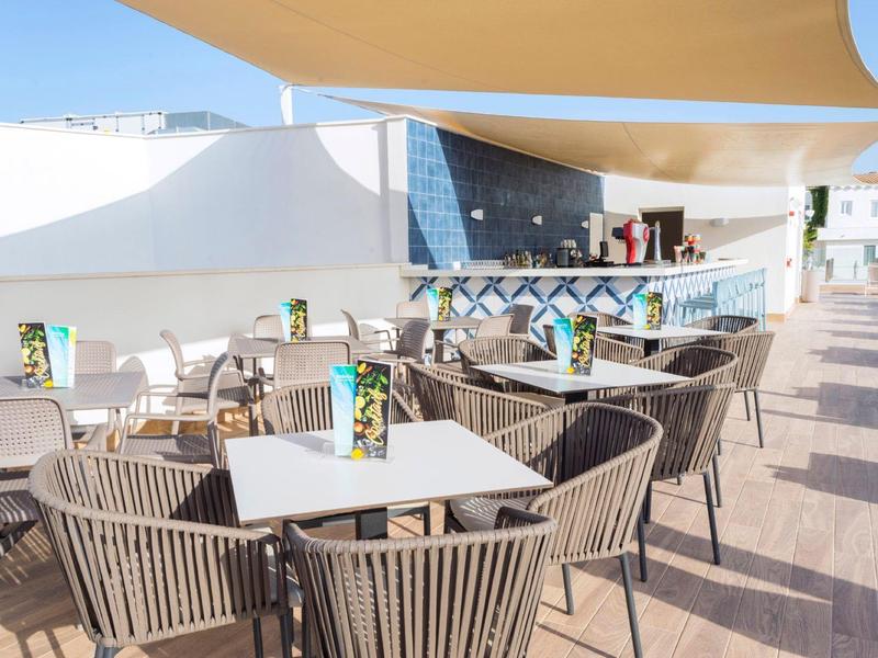 Leerer Außenbereich eines Cafés mit weißen Tischen, grauen Stühlen und Sonnensegeln bei Tageslicht.