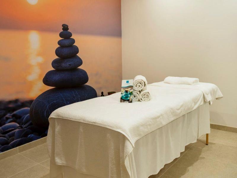 Ruhiger Wellnessraum mit Massageliege, weißer Decke und Wandbild eines Sonnenuntergangs am Meer.