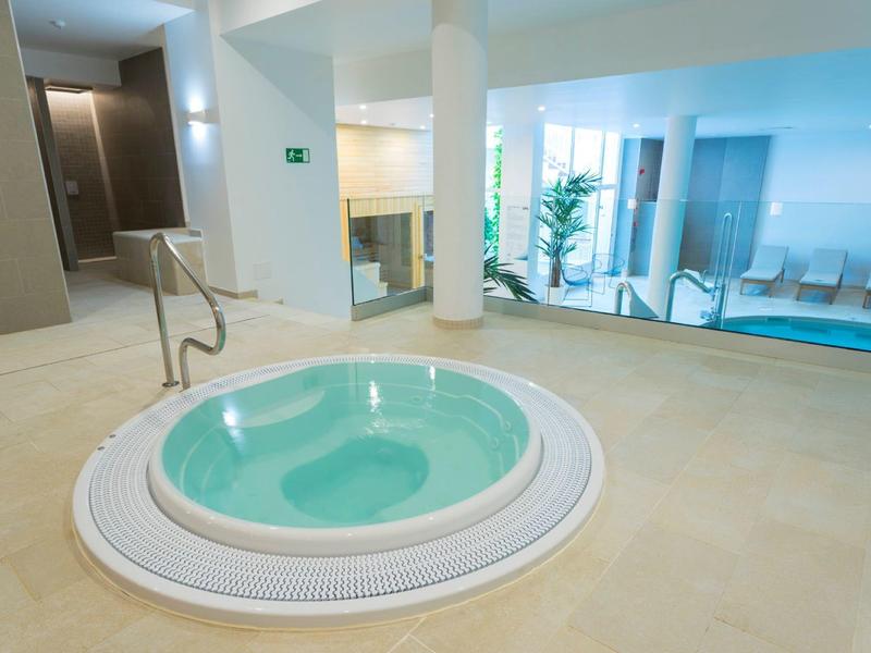 Moderner Wellnessbereich mit rundem Whirlpool, hellem Boden und beleuchtetem Schwimmbecken im Hintergrund.