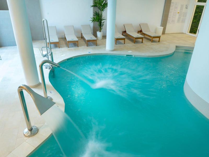 Innenbereich mit blauem Schwimmbad, Wassersprudler, weißen Säulen und mehreren Liegestühlen.