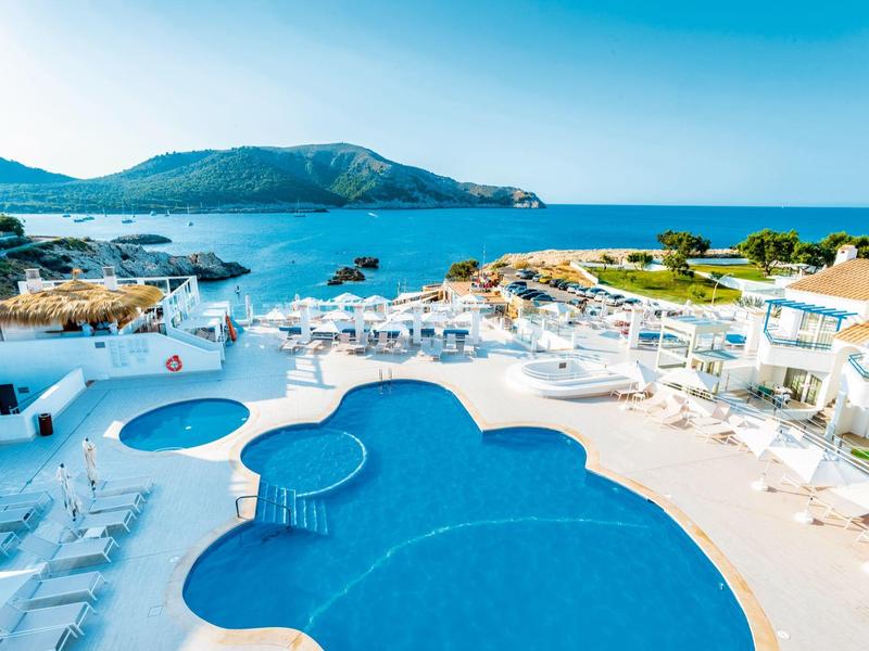 Großer blauer Pool mit Liegestühlen neben dem Meer und grünen Hügeln im Hintergrund.