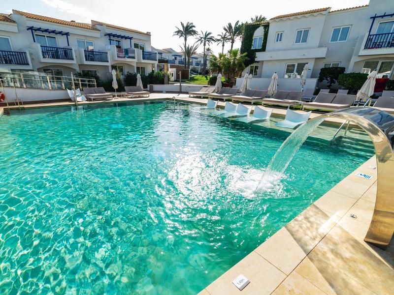 Großer Pool mit klarem Wasser, Wasserfall-Düse, umgeben von Sitzbereichen und Häusern.