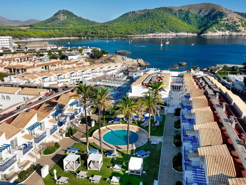 Helle Häuser und Gärten entlang eines Hafens mit blauem Meer und grünen Bergen im Hintergrund.