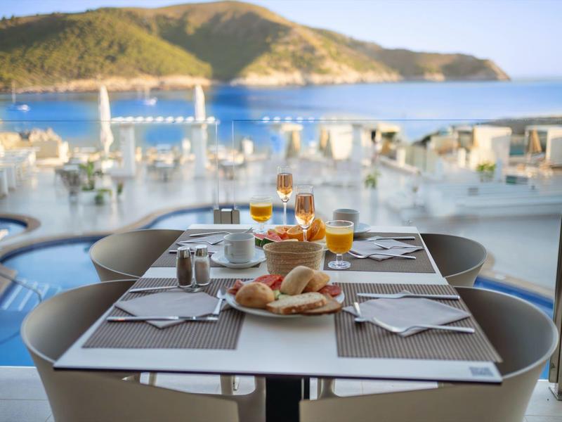 Ein gedeckter Frühstückstisch mit Brötchen, Säften und Kaffee vor Meer und Hügeln.