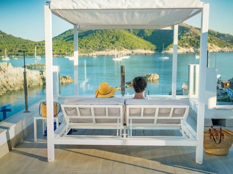 Zwei Personen sitzen auf einer weißen Schaukel vor klarem blauen Wasser und grünen Hügeln.