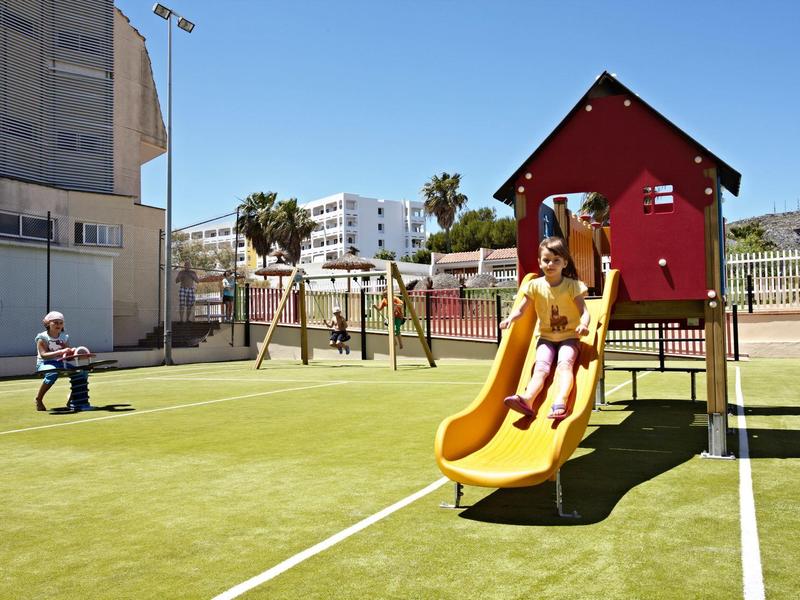 Kind spielt auf gelber Rutsche im Park, andere Person sitzt auf einer Bank, umgeben von Gebäuden.