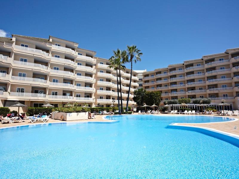 Großer blauer Pool vor einem weißen mehrstöckigen Hotel mit Palmen und Sonnenliegen.