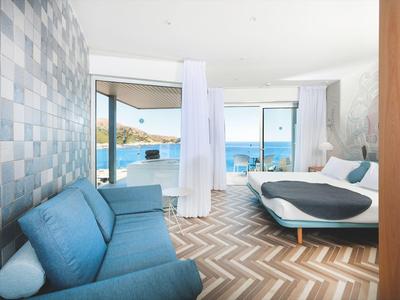 Bright hotel room with blue couch, double bed, and large windows overlooking the sea.