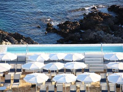 Swimming pool with white loungers and umbrellas next to rocky coast and sea.