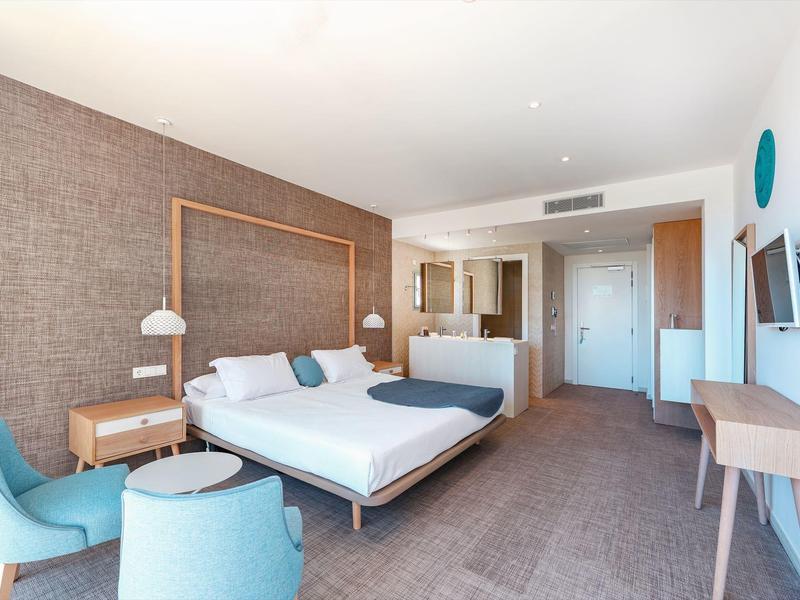 Modern hotel room with large bed, seating area, and desk in neutral tones.