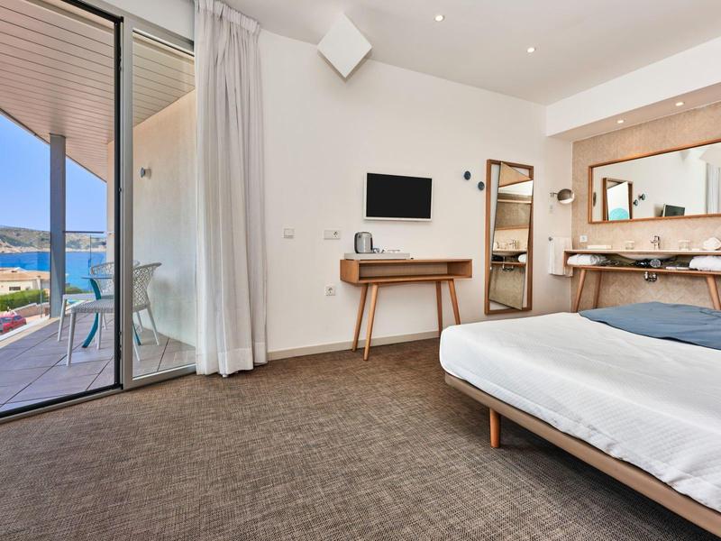 Modern hotel room with double bed, desk, TV, and balcony overlooking the sea.