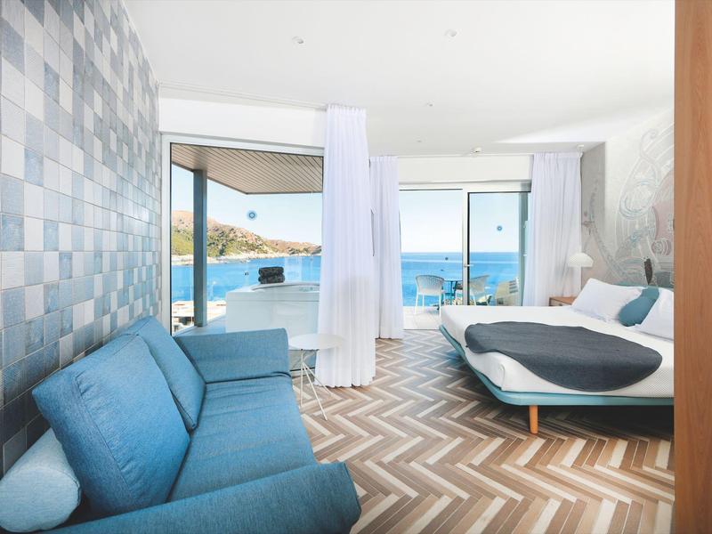 Bright hotel room with blue couch, double bed, and large windows overlooking the sea.