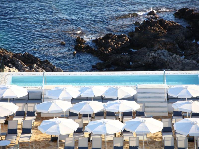 Swimming pool with white loungers and umbrellas next to rocky coast and sea.