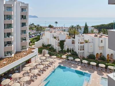 Hotelový komplex s bazénem, lehátky a výhledem na moře v pozadí.