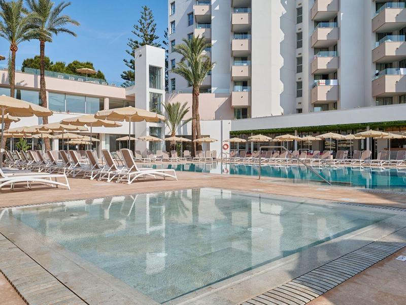 Modern hotel pool area with sun loungers and tall palm trees under a blue sky.