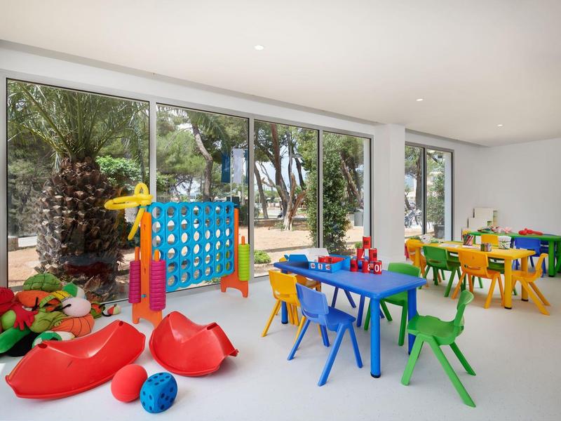 Bright children's playroom with colorful tables, chairs, and toys near large windows with outdoor view.