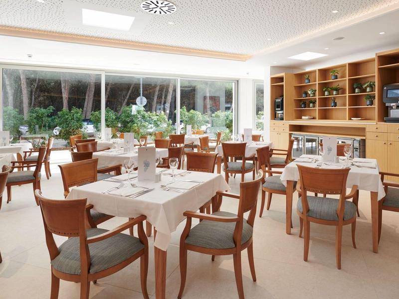 Modern restaurant with wooden tables, chairs, and large windows overlooking greenery.