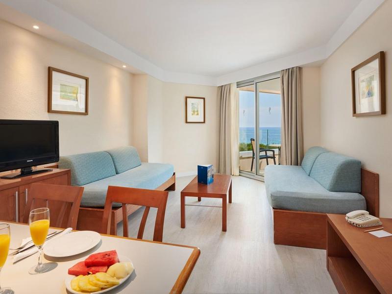 Bright living room with sofas, TV, balcony, and breakfast table with juice and fruit.