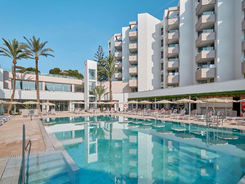 Hotel with large pool, lounge chairs, and palm trees under a clear blue sky.