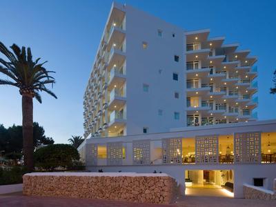 Moderner weißer Hotelbau bei Abenddämmerung mit beleuchteten Balkonen und Palmen im Vordergrund.