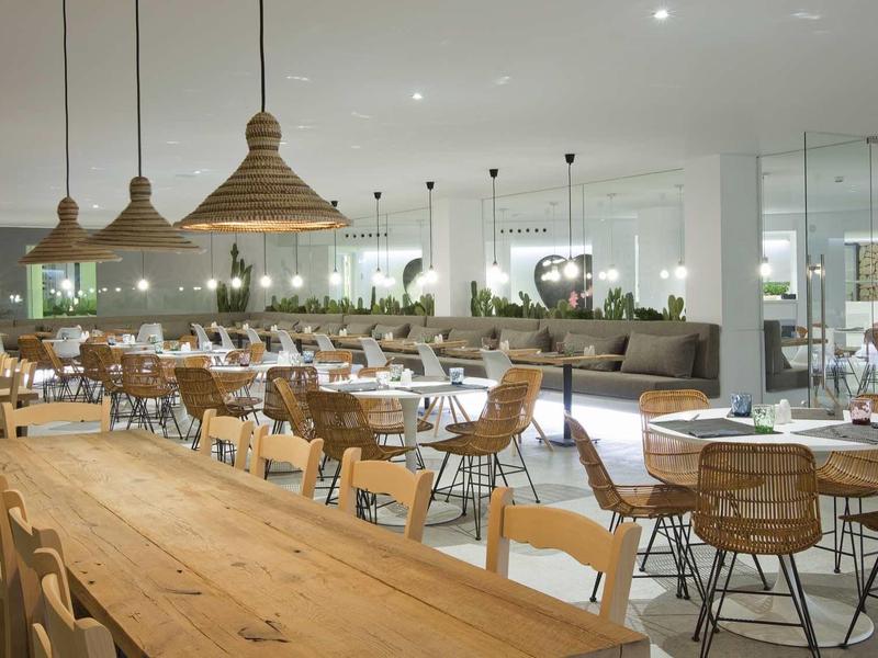 Jasna, nowoczesna restauracja z drewnianymi i rattanymi meblami oraz lampami wiszącymi nad stołami.