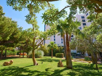 Giardino verde con alberi e statue di fronte a un edificio bianco sotto un cielo soleggiato.