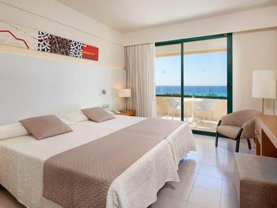 Bright hotel room with double bed, desk, chair, and large window overlooking the sea.