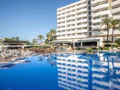 Grande hotel con piscina, palme e ombrelloni sotto un cielo sereno.