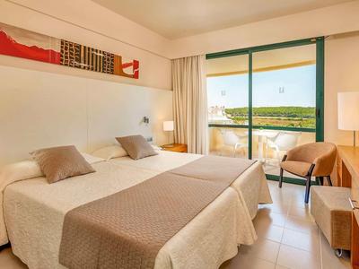 Helles Hotelzimmer mit zwei Einzelbetten, Balkon, Fernseher, Schreibtisch und beigen Dekoelementen.