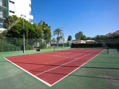 Campo da tennis esterno rosso con linee bianche, circondato da alberi e edifici sotto un cielo sereno.