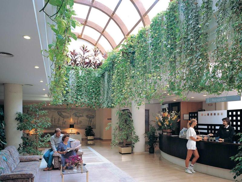 Hotel lobby with green glass roof and guests at the reception and on benches.