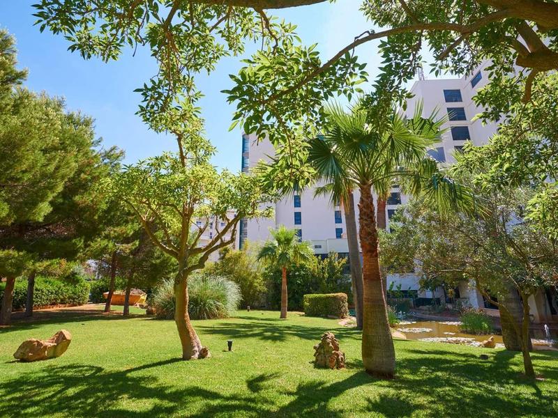 Green garden with trees and grassy areas in front of a white hotel building under blue sky.