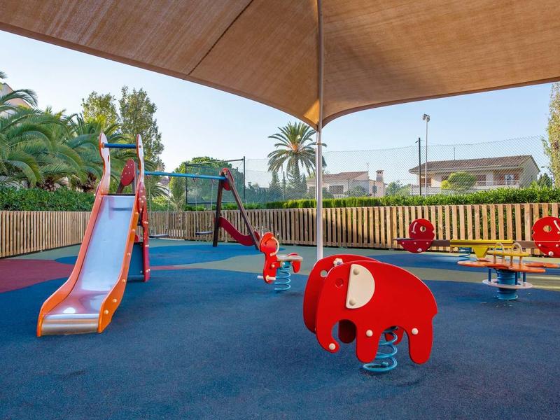 A shaded children's playground with slide, swings, and seesaw under a large sunshade.