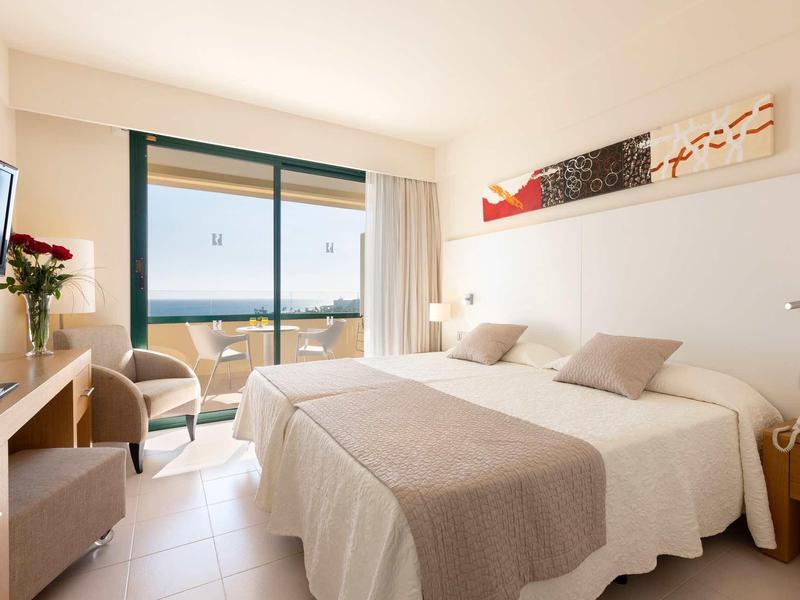 Bright hotel room with two beds, balcony overlooking the sea, and modern interior.