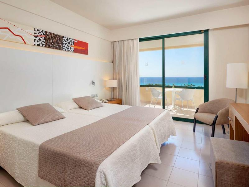Bright hotel room with double bed, desk, chair, and large window overlooking the sea.