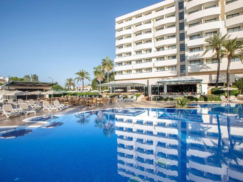 Hotel with large outdoor pool and sun loungers under blue sky.