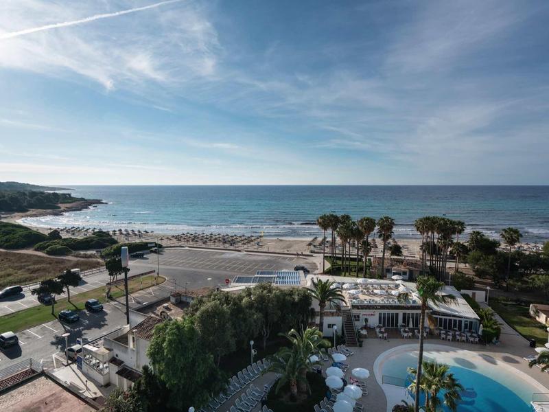 Letecký pohled na pláž se pískem, silnicí, palmami a bazénem pod částečně zamračenou oblohou.