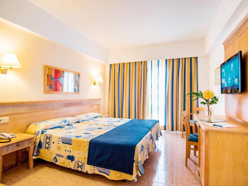 Bright hotel room with double bed, wooden furniture, and yellow-blue curtains.
