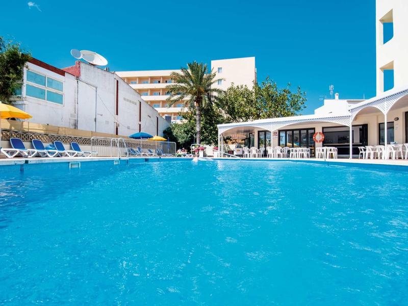 Large blue hotel pool with sun loungers and buildings in the background under clear sky.