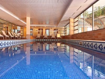 Helles Hallenbad mit blauen Fliesen, Glasfenstern und Sitzbereich im warmen Holzdesign.