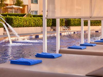 Poolbereich mit Liegestühlen, blauen Handtüchern und Wasserspielen an einem Hotel.
