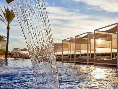 Wasserfall strömt in Pool mit Palmengarten und weißen Lounge-Betten bei Sonnenuntergang.