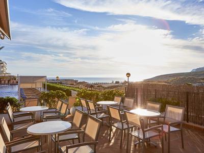 Terrassenrestaurant mit Tischen und Stühlen, Blick auf Meer und Hügel bei Sonnenuntergang.