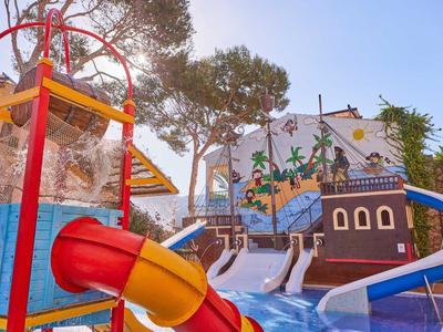 Buntes Wasserspielplatz mit Rutschen, Wasserfall und Piratenschiff-Motiv unter Bäumen bei Sonnenschein.